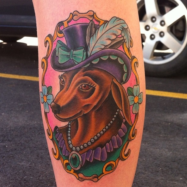 artistic Dachshund wearing hat with ribbon and feathers, pearl necklace around its neck in a vintage gold frame tattoo on the leg