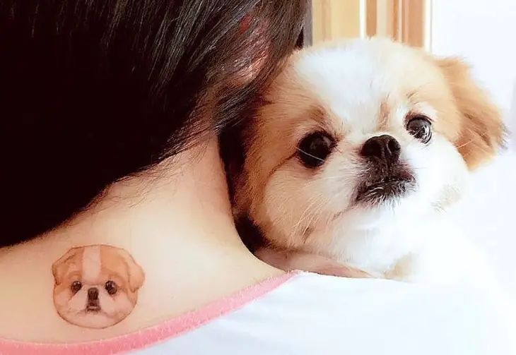 a woman carrying her Pekingese dog while showing her Pekingese tattoo on the back of her neck