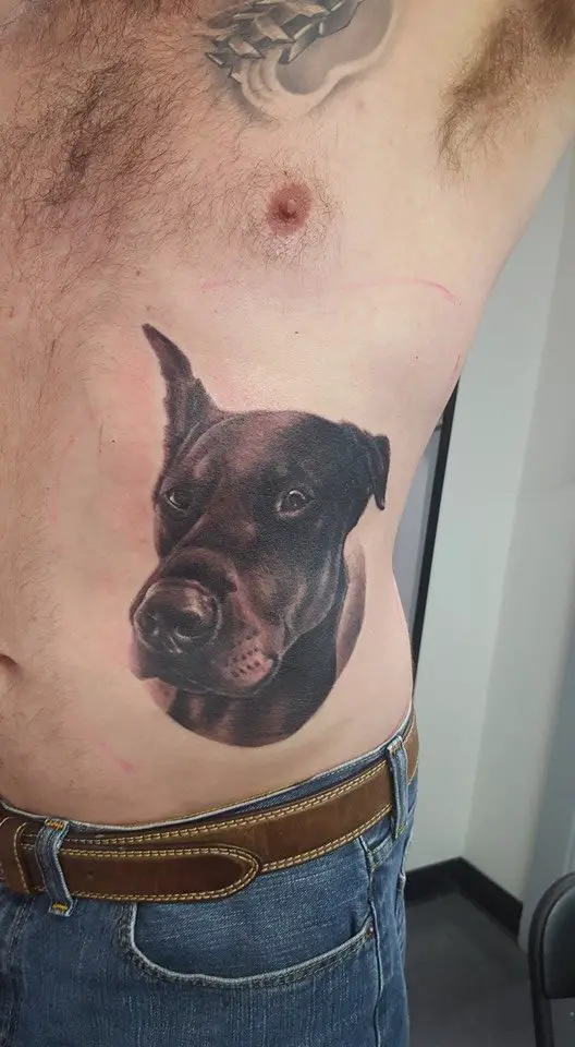 black and gray face of a Doberman with its one ear up tattoo on the side of a man's body