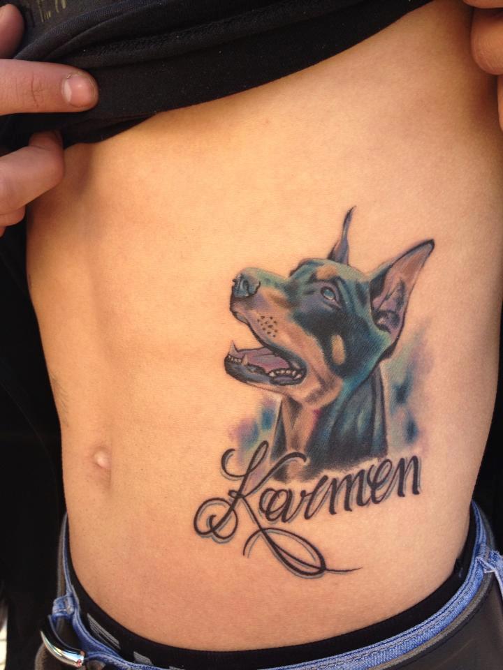 looking up Doberman while smiling and with name - Karmen tattoo on the side of a man's body