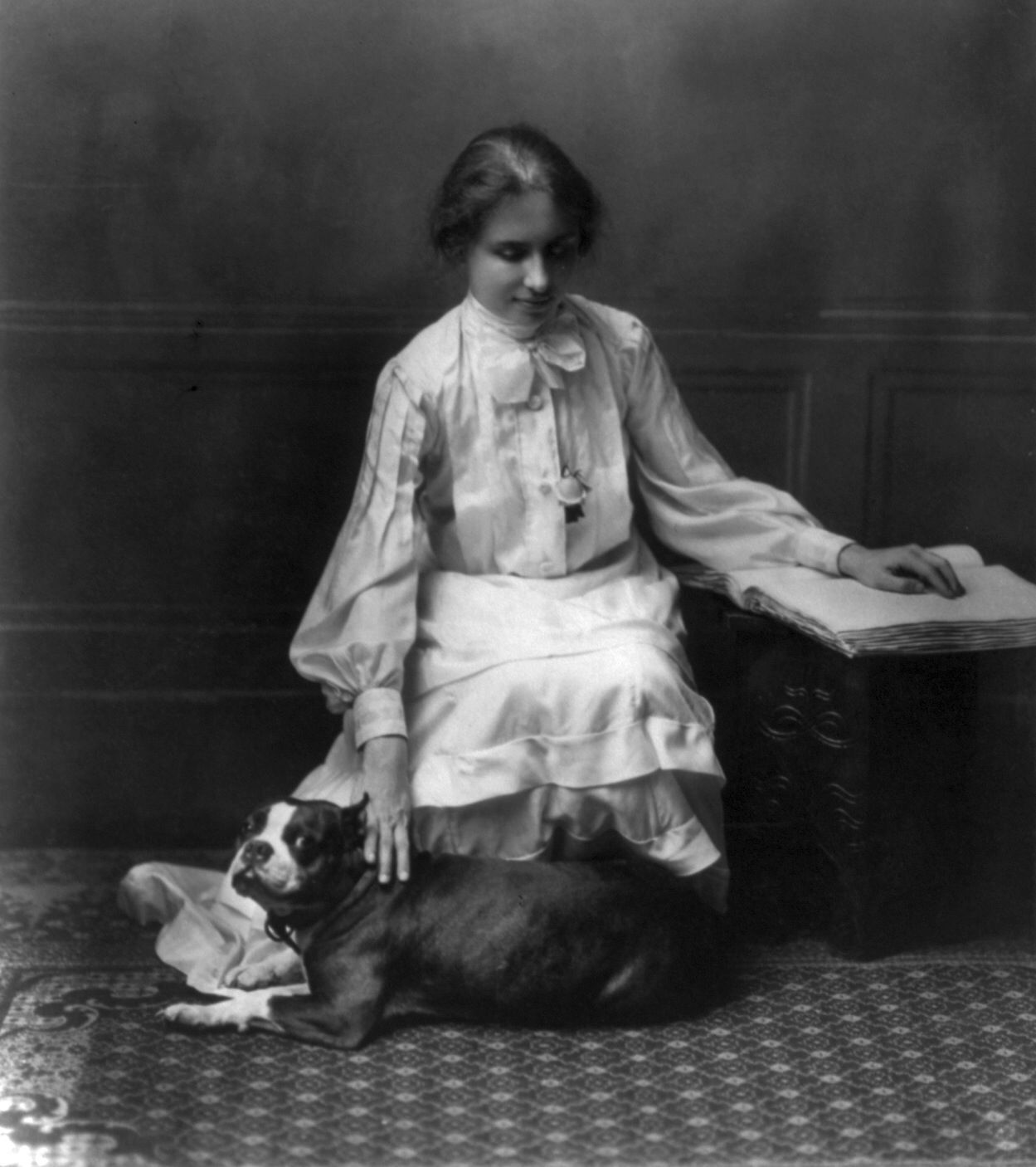 Helen Keller sitting on the chair while touching the back of her Boston Terrier lying on the floor on her feet