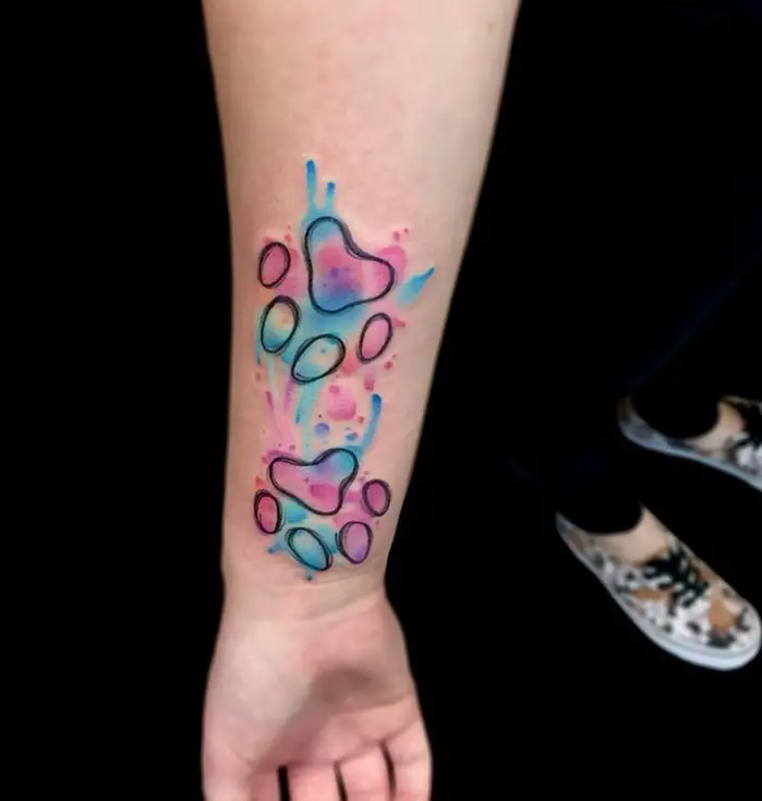 outline of paw prints with colorful splashes of watercolor tattoo on the forearm