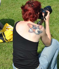 a corgi face tattoo on the back of a woman sitting on the grass while taking a photo using her camera