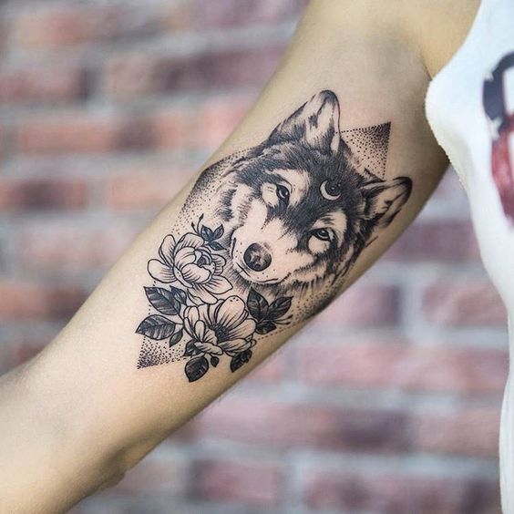 face of a Husky with crescent moon on its forehead and with flowers underneath him inside a dotted diamond shape tattoo on the biceps of a woman