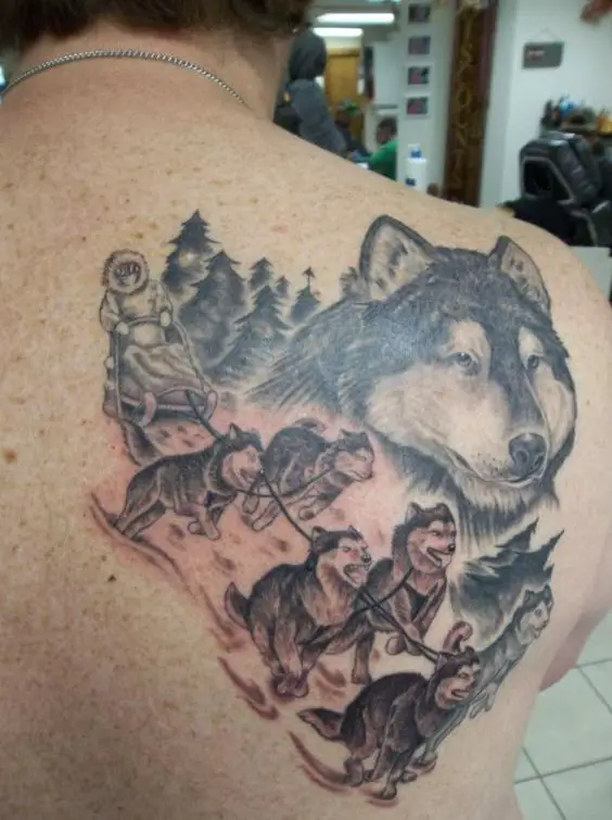 a man riding a pack of Husky sled in snow with a large face of Husky next to them tattoo on the back