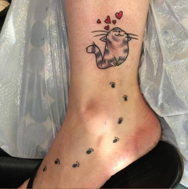 a smiling cat with red hearts on top of its head and paw prints below it tattoo in the ankle going to the foot of the woman