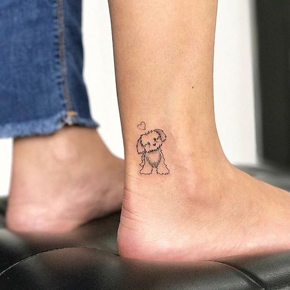 outline tattoo of a yorkie on the ankle of a woman standing on top of the chair