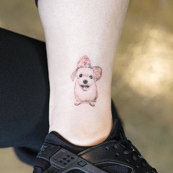 small white dog wearing a colorful big ribbon on its head tattoo on the ankle