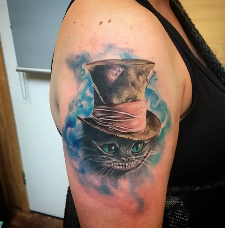 black Cheshire Cat with green eyes wearing a brown hat wrapped with pink fabric in a blue and green watercolor background tattoo on the shoulder