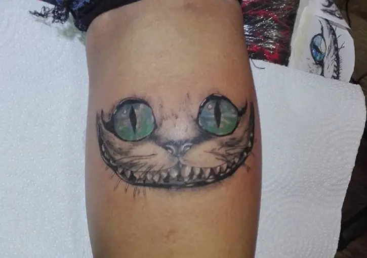 black and gray Cheshire Cat with green and blue eyes tattoo on the leg