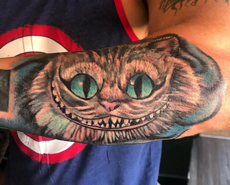 Cheshire Cat with pink nose and aqua blue eyes in a blue shadow background tattoo on the arm