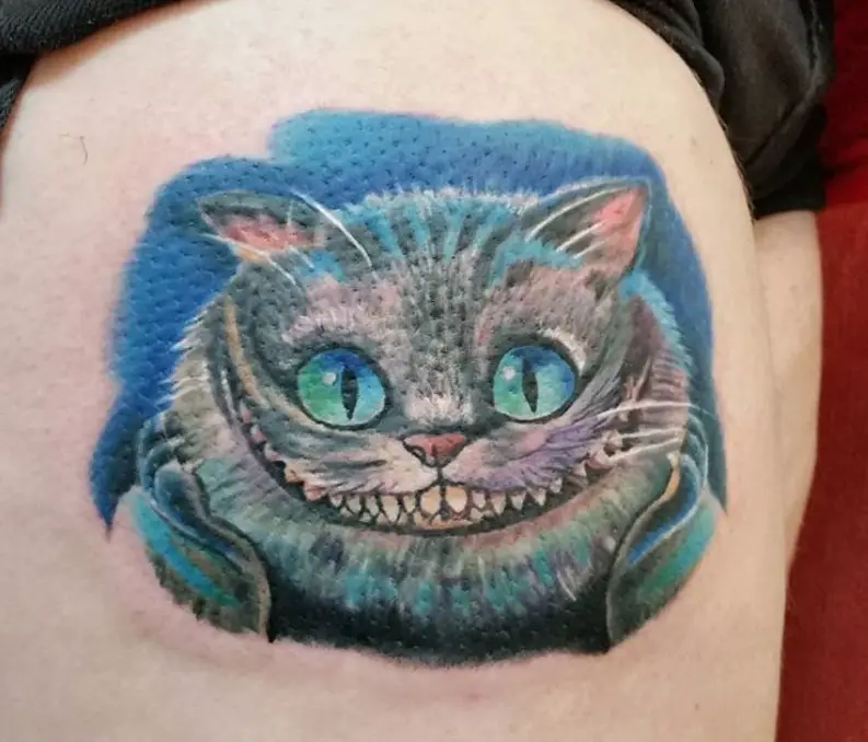gray Cheshire Cat with hints of blue, purple, and green colors in its fur and blue and green eyes, in a blue shadow tattoo on the thigh