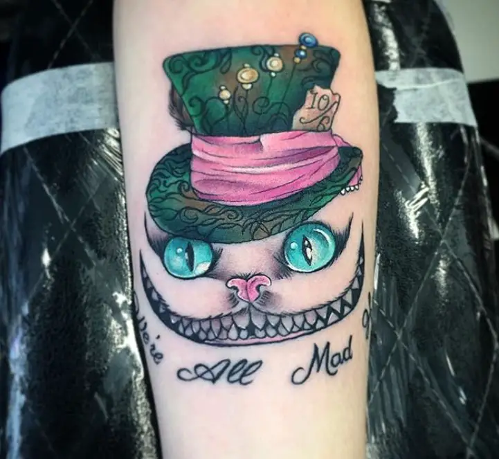black and gray Cheshire Cat with pink nose and aqua blue eyes wearing a green hat with wrapped with pink ribbon tattoo on the forearm