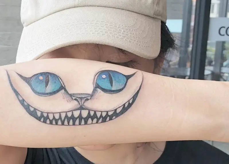 a lady showing her forearm with a tattoo of a Cheshire Cat with blue eyes