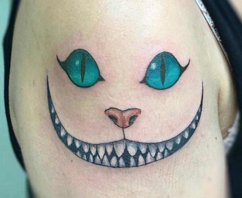 simple Cheshire Cat smiling with its white teeth, red nose, and blue green eyes tattoo on the shoulder