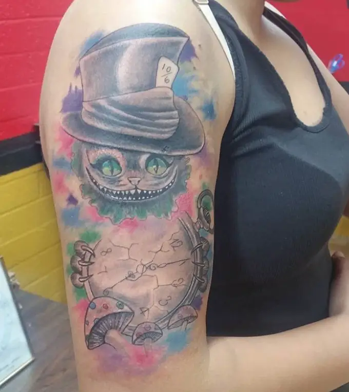 gray Cheshire Cat with blue eyes wearing a hat behind a timer clock and surrounded with splashes of colors tattoo on the shoulder