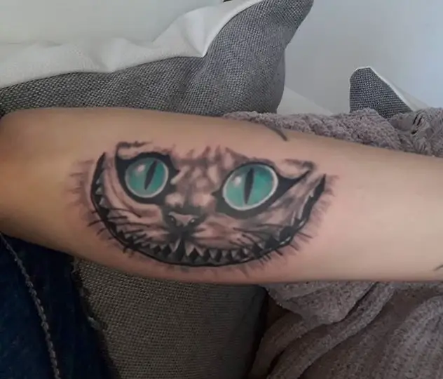 black and gray Cheshire Cat with aqua blue eyes tattoo on the forearm