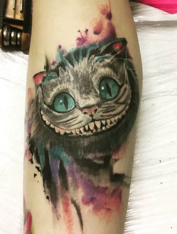 gray Cheshire Cat with green eyes and in pink, purple and blue watercolor background tattoo on the leg