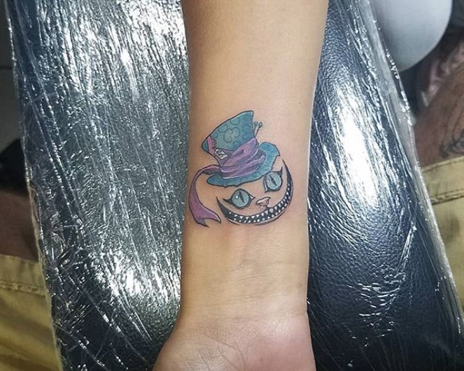 Cheshire Cat with blue eyes wearing a blue hat with wrapped with purple fabric tattoo on the forearm