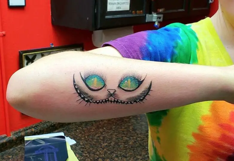 black and gray Cheshire Cat with blue, green, yellow, and pink eyes tattoo on the arm