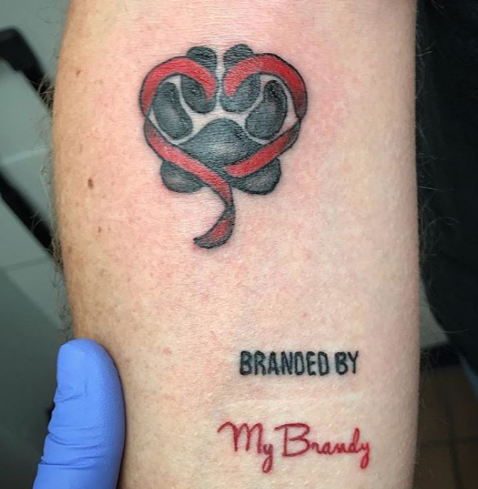 red heart ribbon with black paw print behind tattoo on forearm