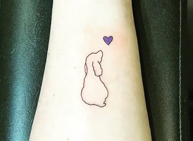 outline of sitting dog looking at the small purple heart tattoo on forearm