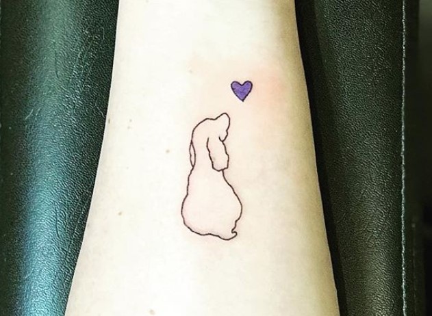 outline of sitting dog looking at the small purple heart tattoo on forearm