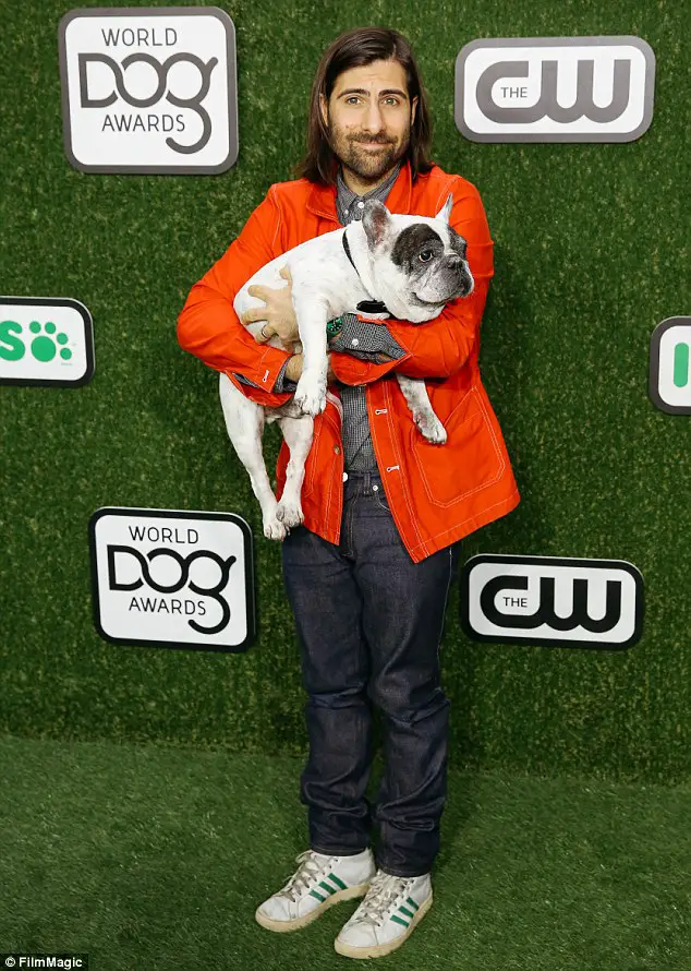 Jason Schwartzman standing while carrying his adult French Bulldog