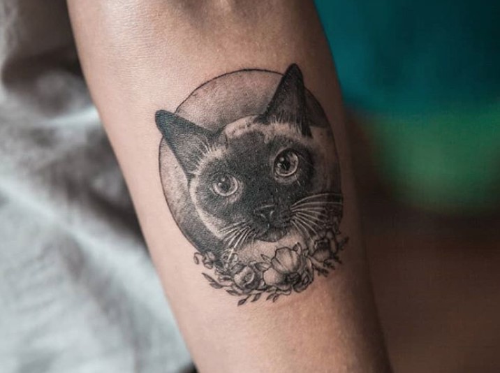 forearm of a person tattooed with an adorable face of a Siamese Cat inside a circle shape with flowers on the bottom