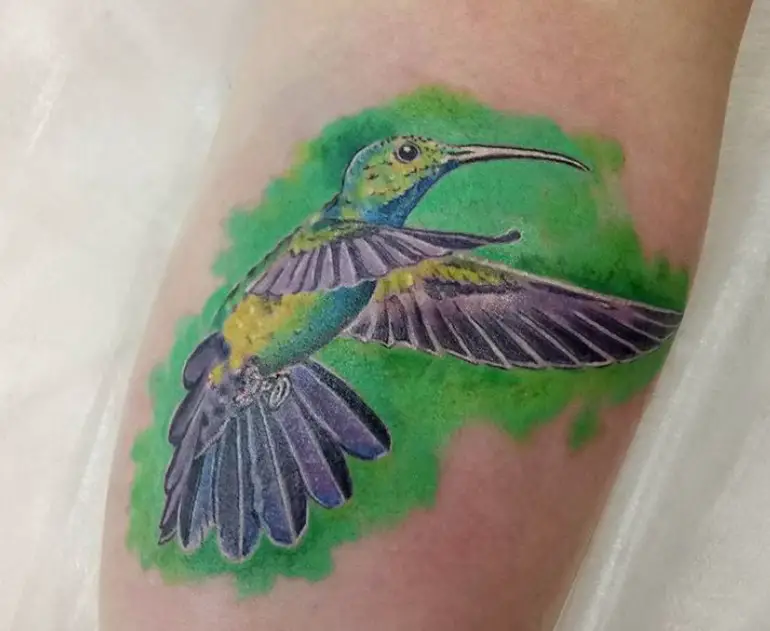 violet, blue, and yellow colored hummingbird with green watercolor tattoo on forearm