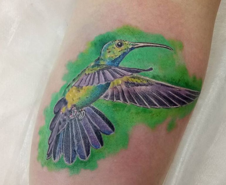 violet, blue, and yellow colored hummingbird with green watercolor tattoo on forearm