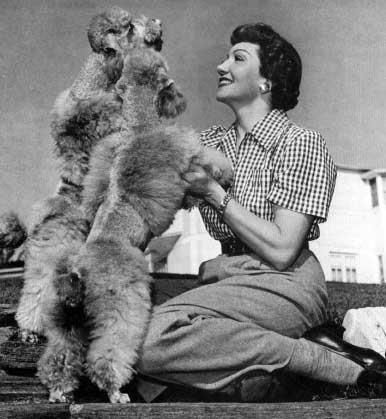 Claudette Colbert sitting in the yard while holding the arms of her poodle standing in front of her