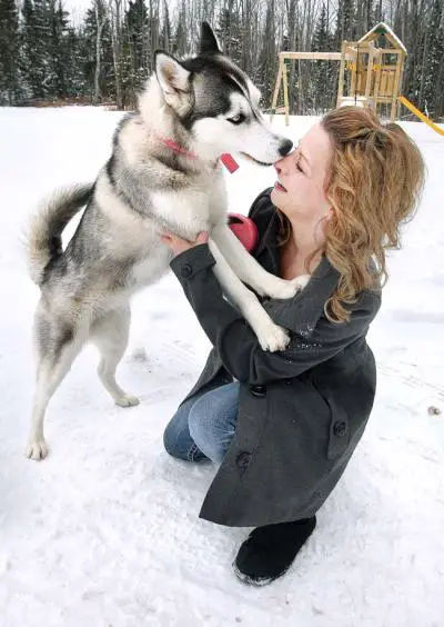 A woman kneeling in snow while being kissed in the nose by a Husky