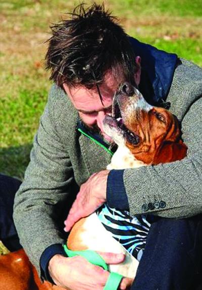 David Arquette sitting on the grass while hugging a Basset Hound