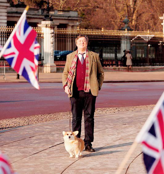 Stephen Fry walking in the street with his Corgi behind the small flags of USA 