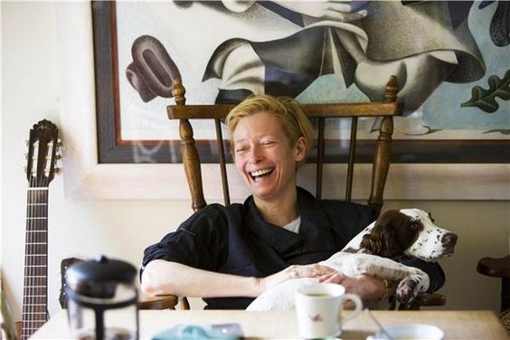 Tilda Swinton laughing while sitting on the chair with her Springer Spaniel in her lap
