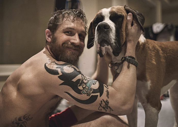 Tom Hardy sitting on the floor while petting his Boxer Dog standing in front of him