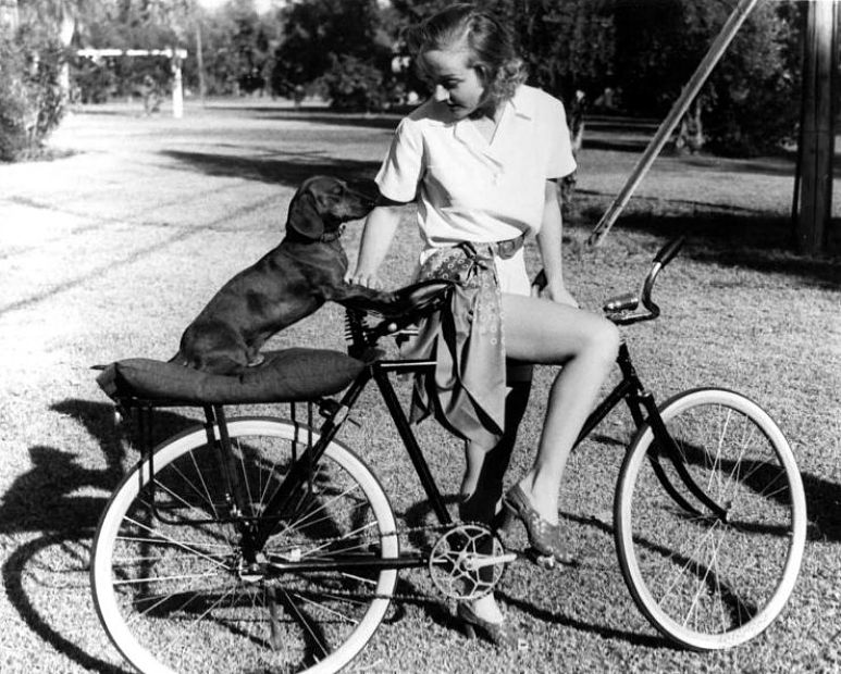 Bridgitte Bardot riding a bike while her Dachshund is sitting on the seat behind her