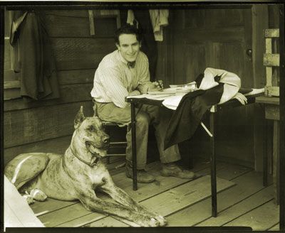 Harold Lloyd sitting in the table with his Great Dane lying on the floor