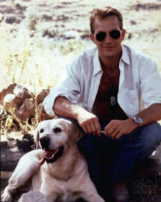 Kevin Costner sitting on a laid tree trunk with his Labrador sitting underneath him