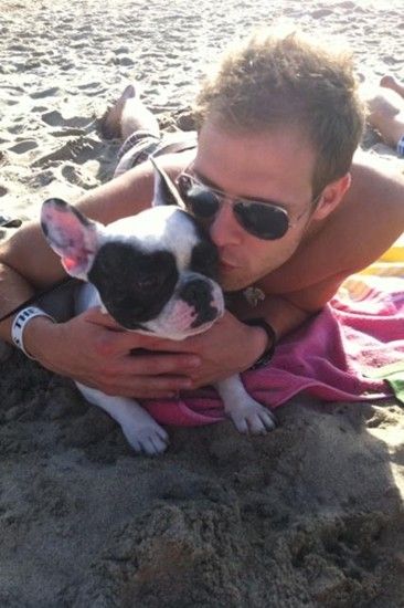 Kris Allen lying down in the sand while kissing the cheeks of his French Bulldog