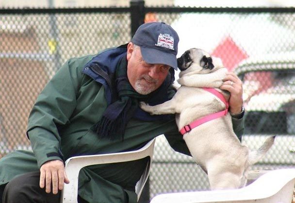 Billy Joel sitting on the chair while hugging his pug standing up from the chair behind him