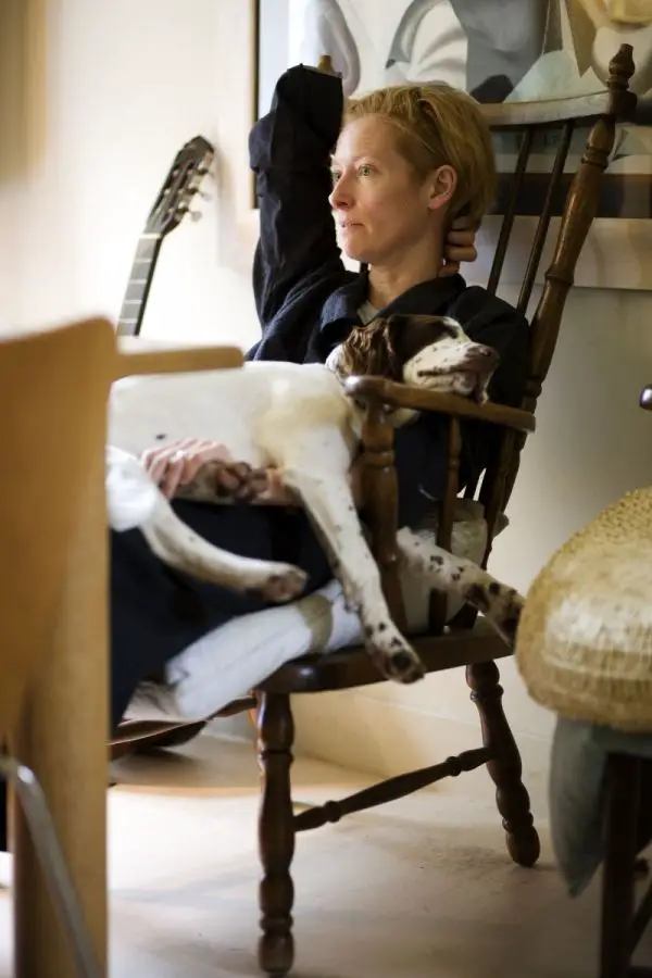 Tilda Swinton sitting on a chair while her Springer Spaniel dog is sleeping on her lap