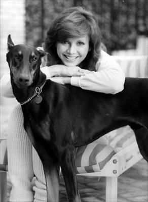 Victoria Principal sitting on the chair with her head and arms on top of the back of a Doberman standing in front of her