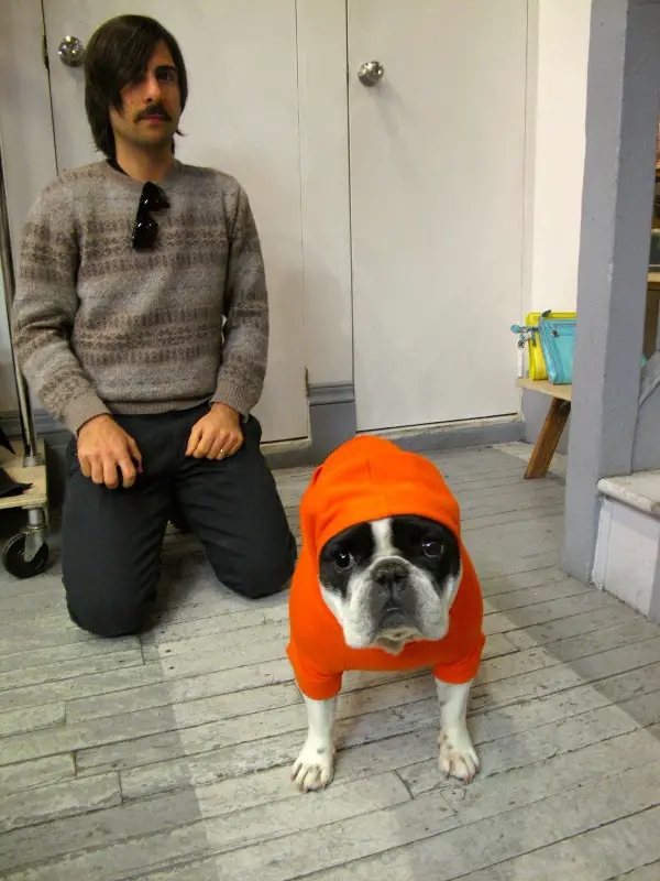 Jason Schwartzman kneeling behind his French Bulldog standing on the floor while wearing an orange jacket