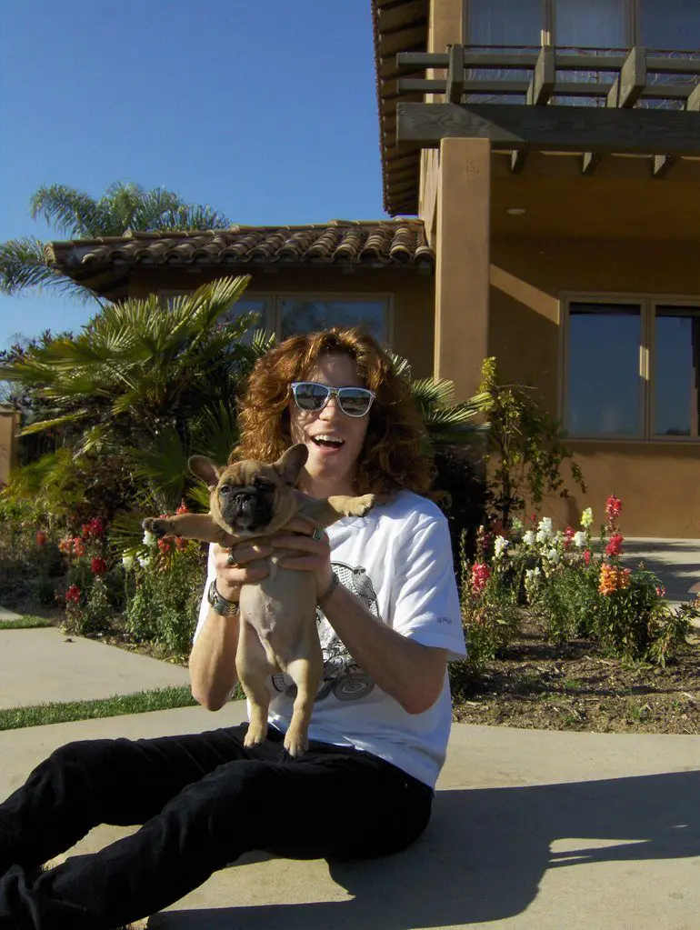 A Shaun White sitting on the pavement while holding up his French Bulldog puppy