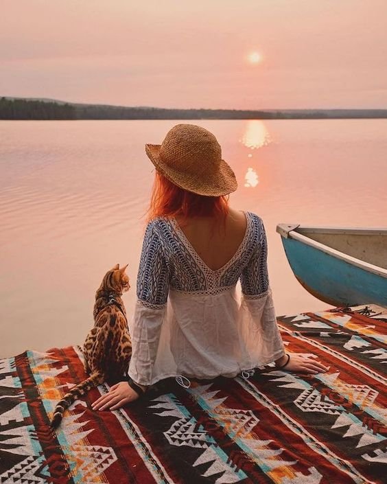 A Bengal Cat sitting next to a woman facing the view of the ocean on a sunset