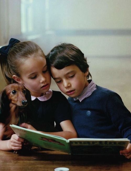 a girl with a Dachshund sitting beside her and boy at the table reading a book