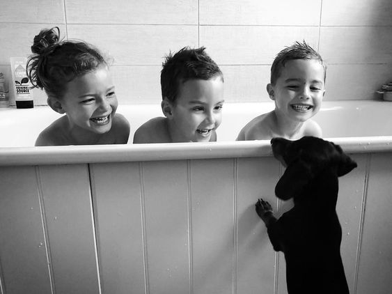 three happy children in the bathtub while a dachshund is looking at them