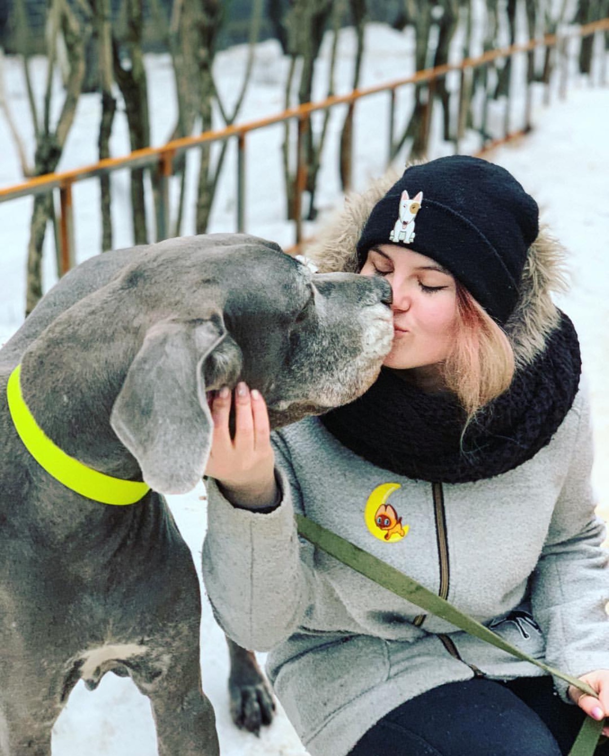 A woman outdoors in snow while kissing the mouth of a great dane standing beside her
